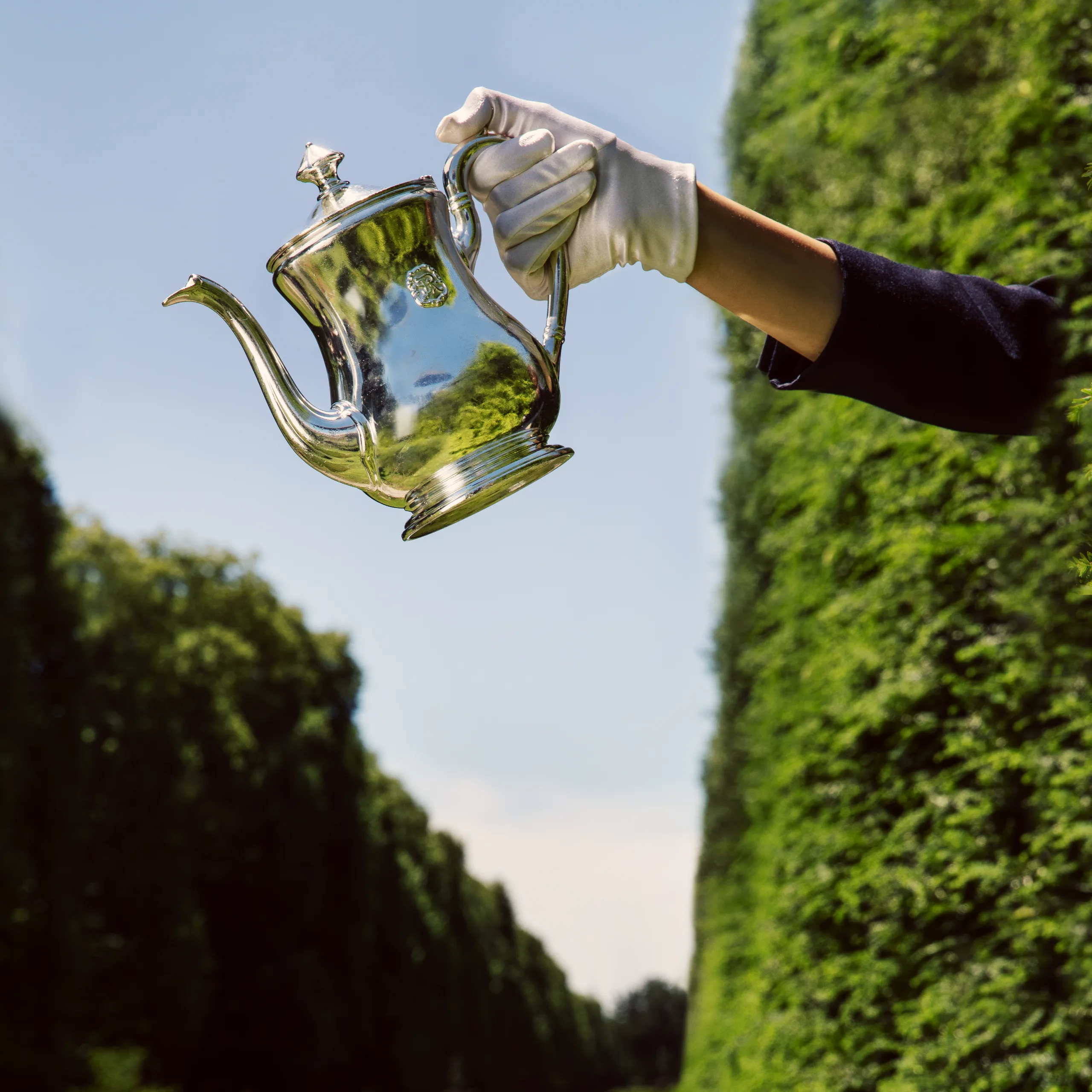 A gloved hand holding a shiny silver teapot against a clear blue sky and green trees