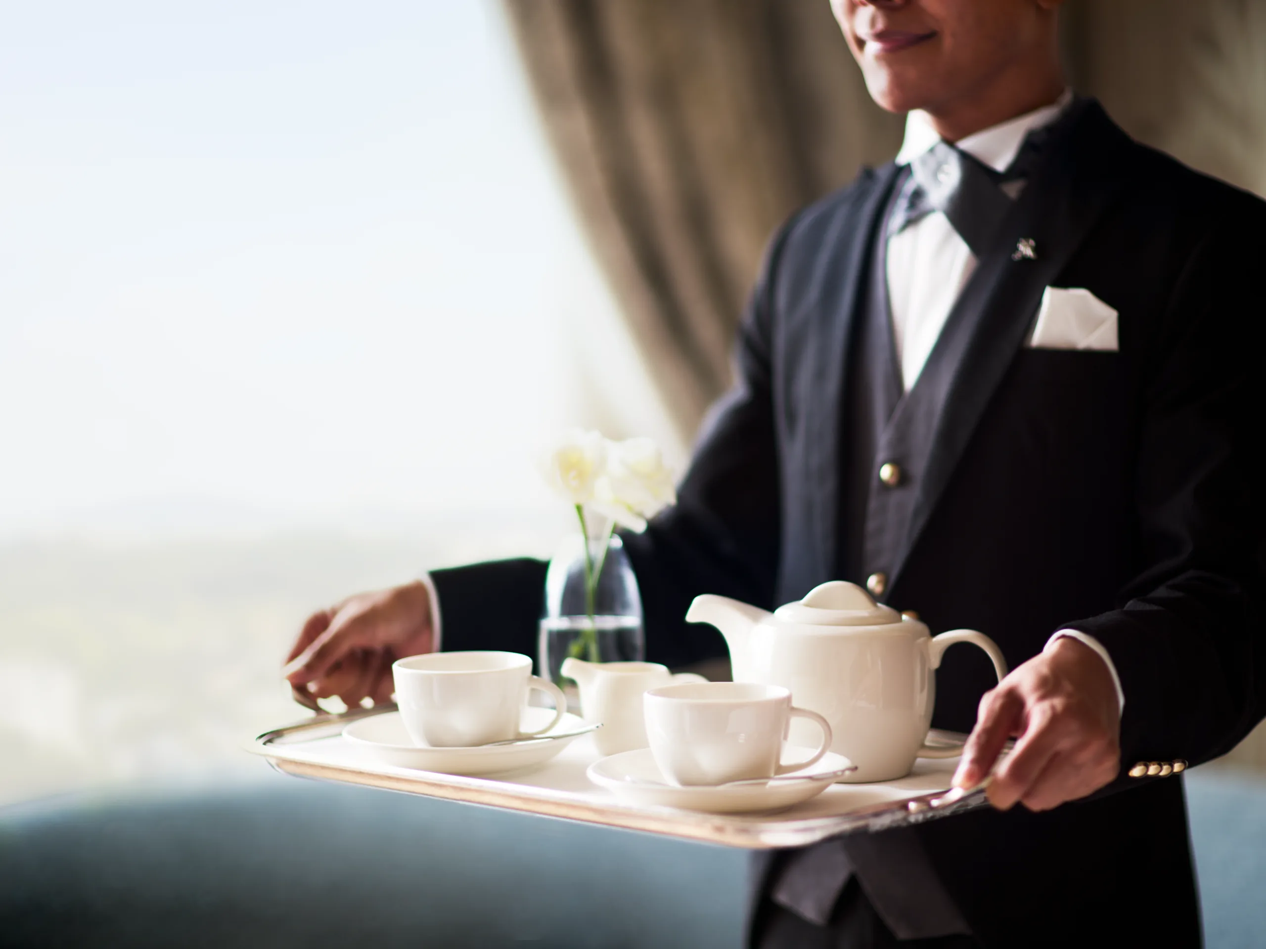 A person in formal attire serving tea with a tray of white teapots and cups