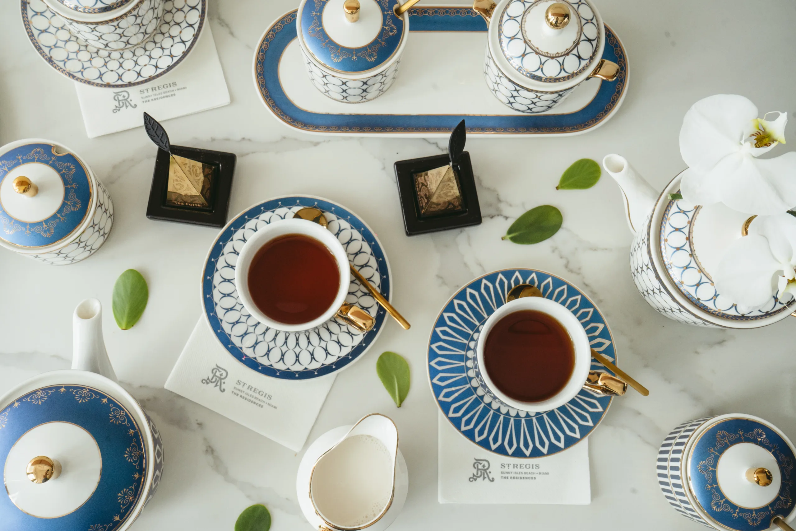 Blue and white porcelain tea set with gold accents on a marble table, with tea cups filled with tea and green leaves scattered around